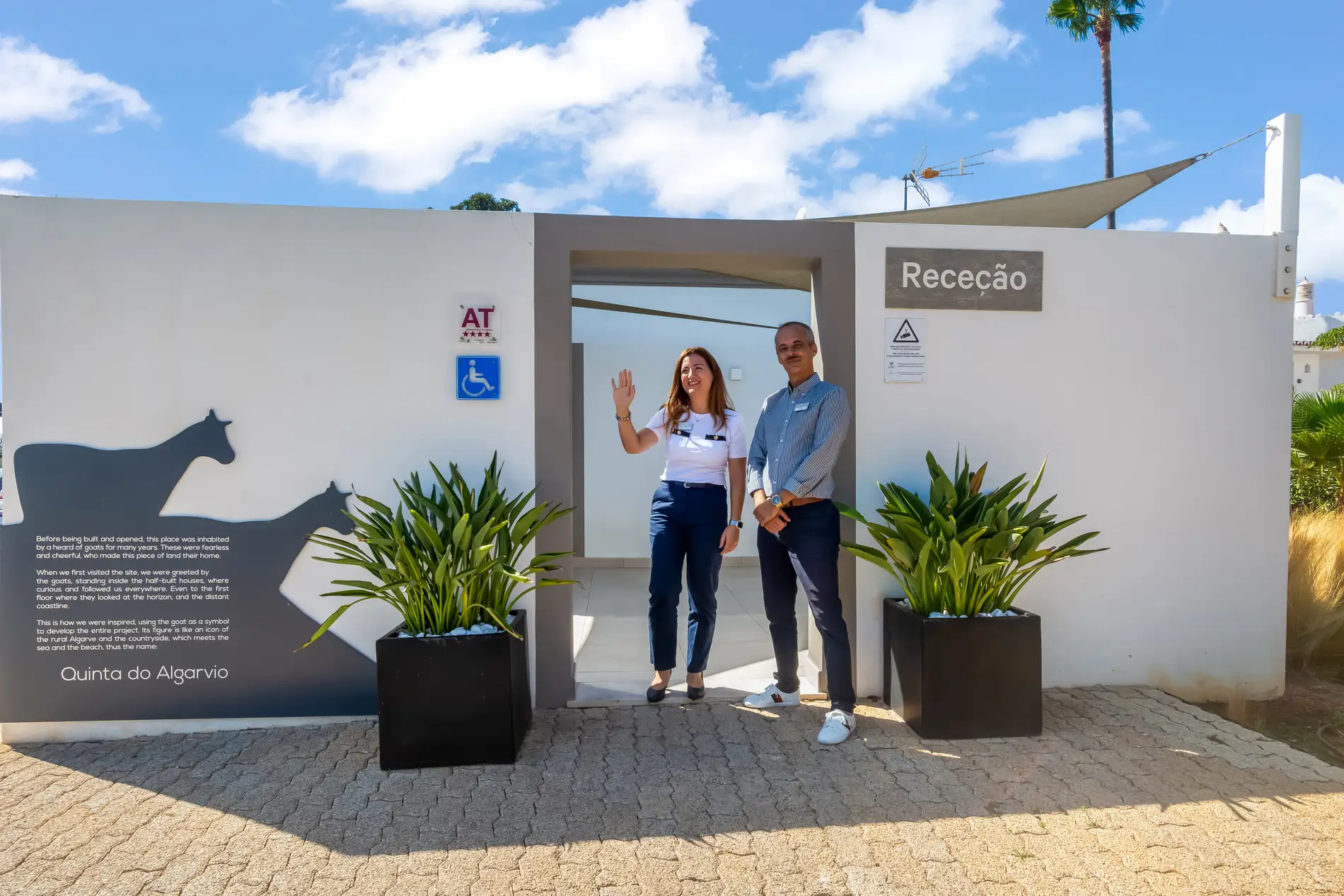 Receptionists at a modern entrance with potted plants and a blue sky.