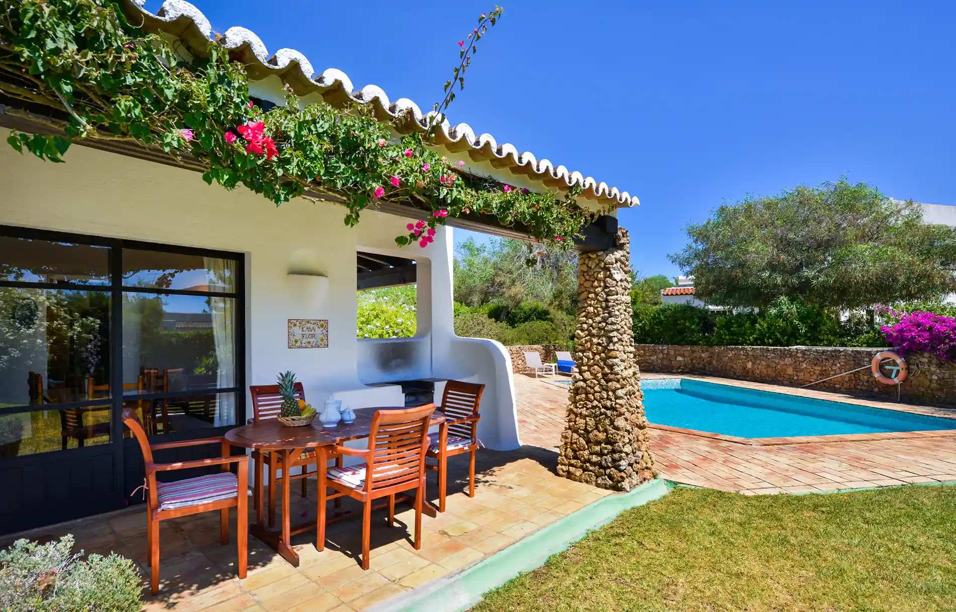 Covered patio with dining set beside a pool, surrounded by greenery.