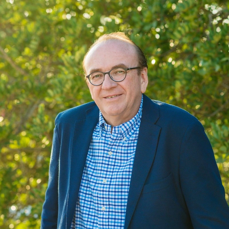 Man in blue blazer smiles outdoors with leafy background.