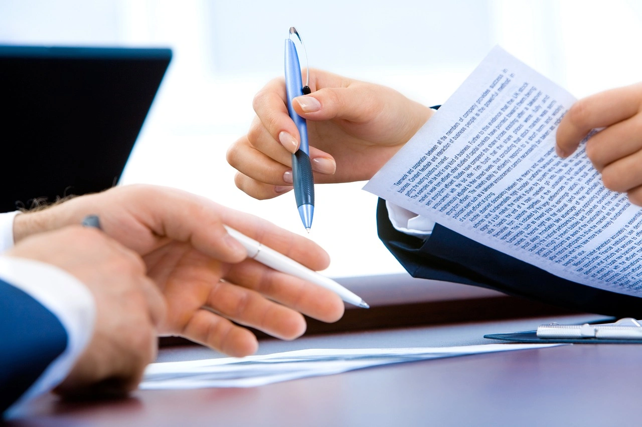 Hands exchanging pens and documents over a table in an office setting.