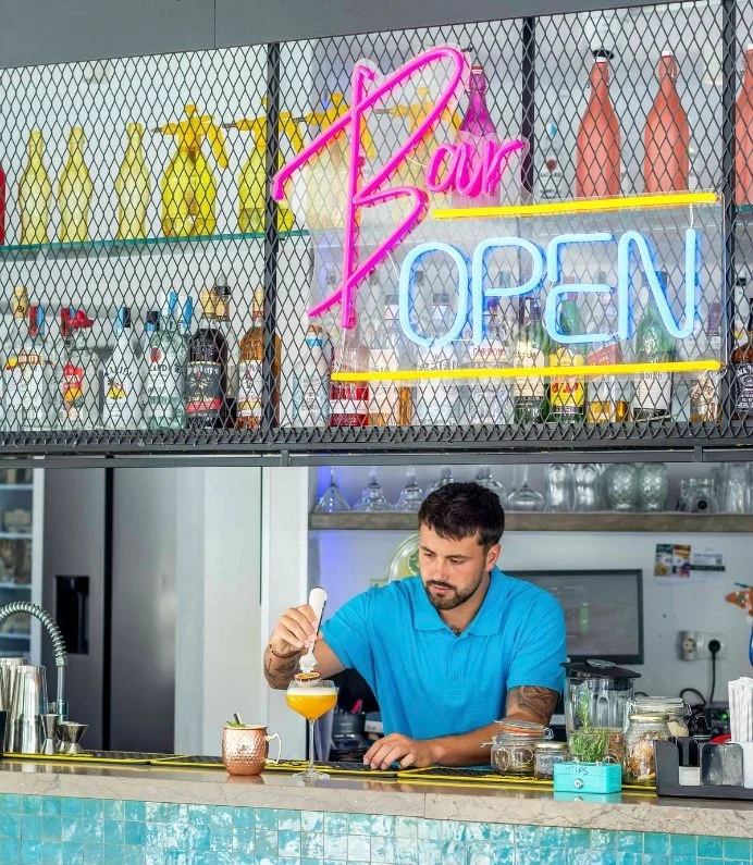 Bartender preparing a cocktail under a "Bar Open" neon sign.