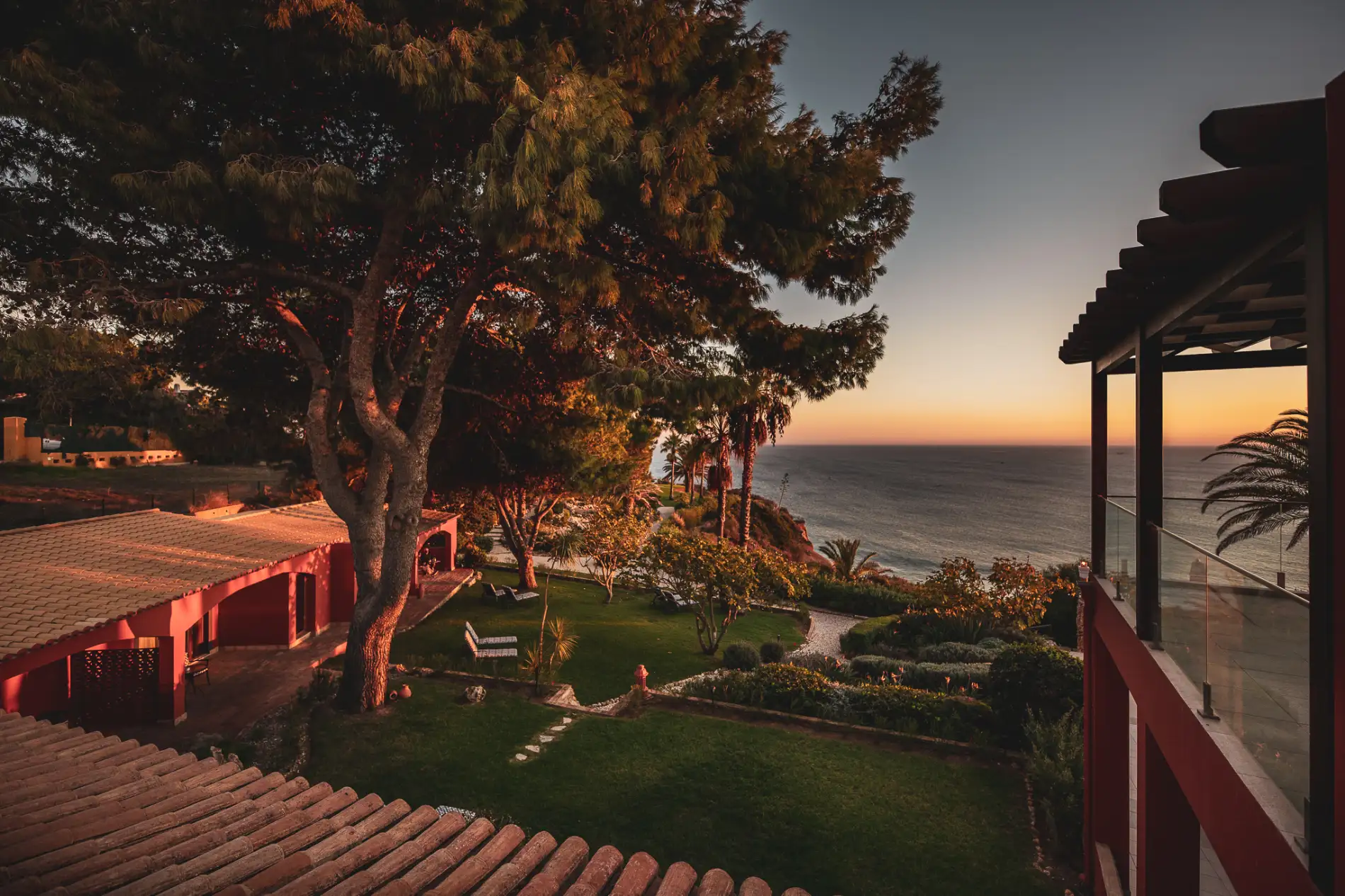 Sunset view over ocean from a garden with trees and house.