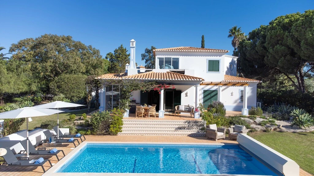 White villa with pool in a lush, sunny garden setting.