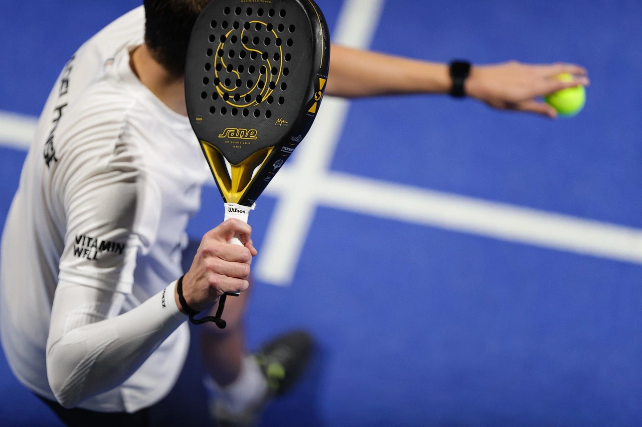 Close-up of person holding a padel racket and ball on a blue court.
