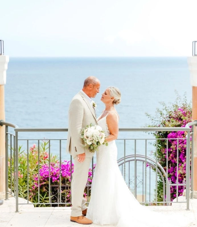 Bride and groom embrace by the sea, surrounded by vibrant flowers.