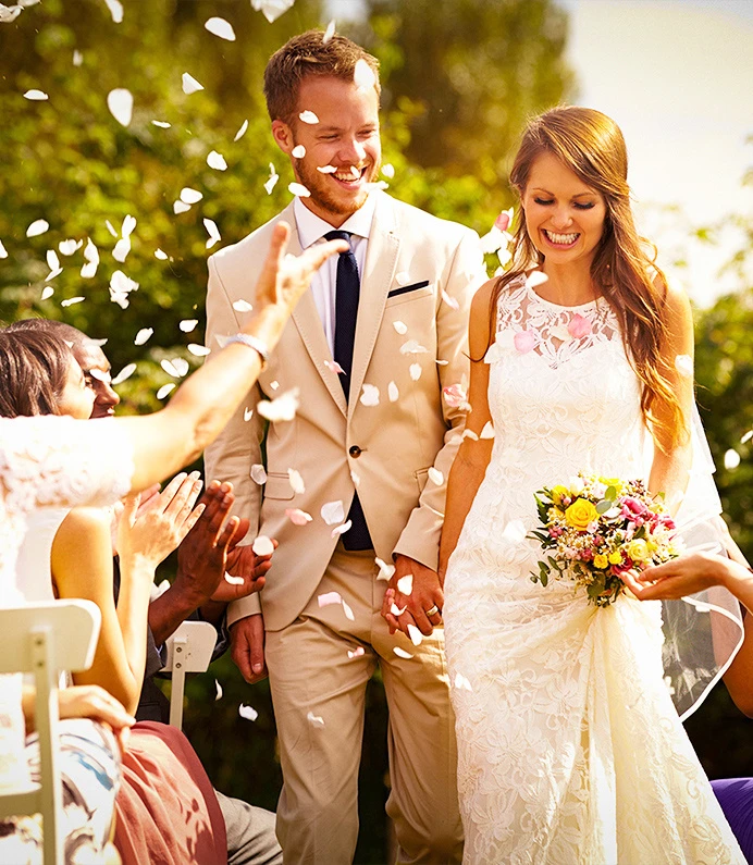 Bride and groom smile amid flower petals at an outdoor wedding ceremony.