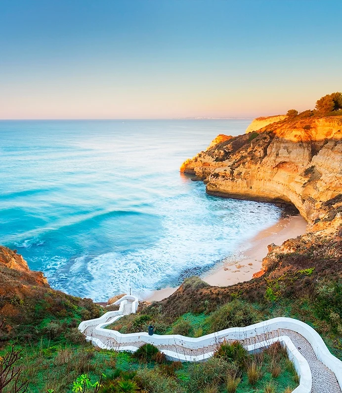 Path leads to a beach cove at sunrise, with cliffs and blue sea.