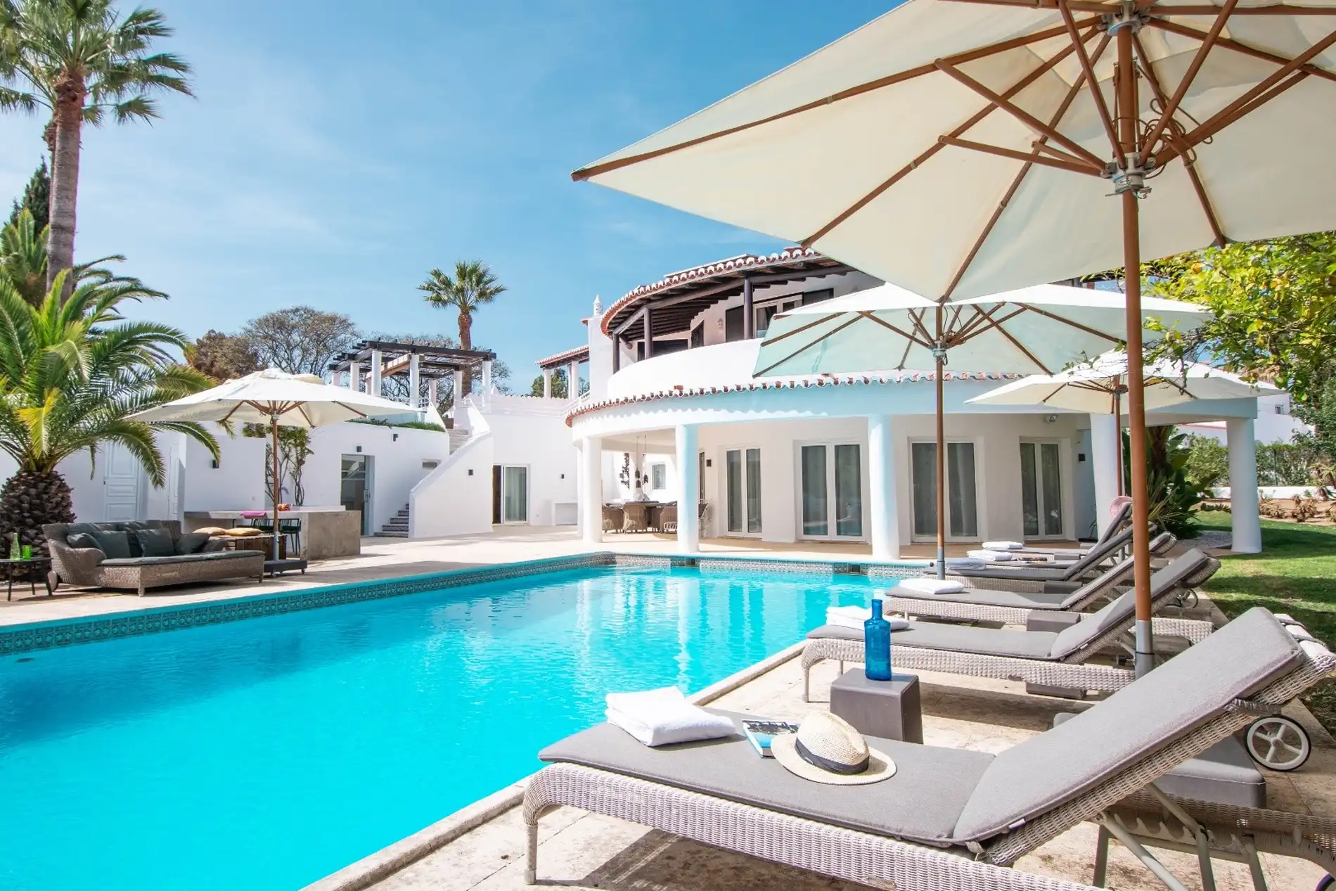 Luxurious villa with pool, lounge chairs, and umbrellas under a clear blue sky.