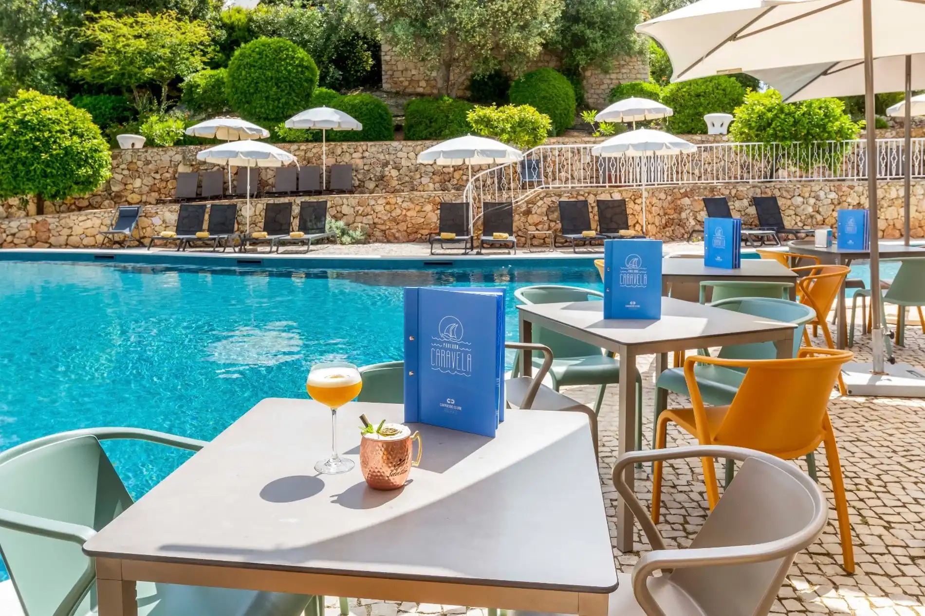 Poolside chairs and tables with drinks under umbrellas on a sunny day.