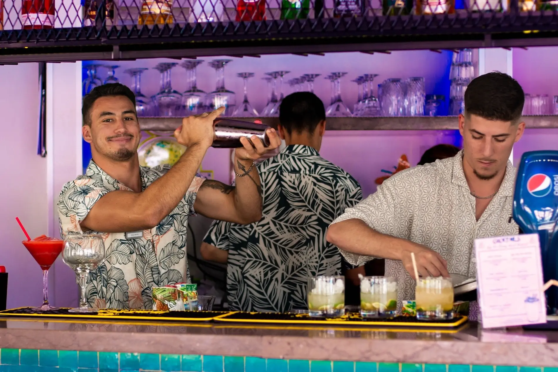 Bartenders making drinks behind a colorful bar decorated with glasses.