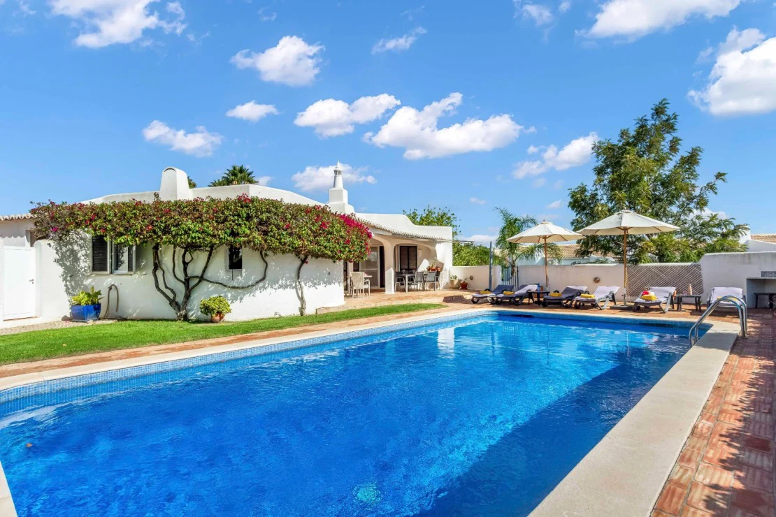 White villa with pool, sun loungers, and umbrella under a blue sky.