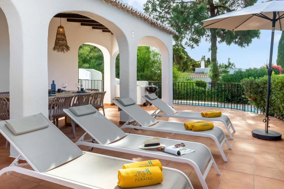 Poolside loungers with sunhats under umbrella, near patio dining area.