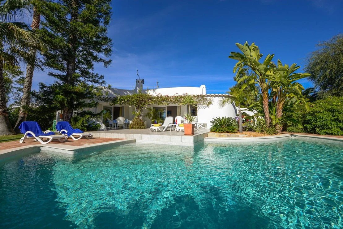 House with pool, sun loungers, and lush greenery under a clear blue sky.
