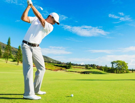 Golfer in white swings club on sunny golf course with blue sky.