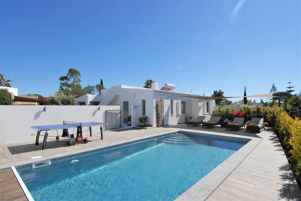 Modern villa with pool, sun loungers, and a ping pong table under a clear sky.