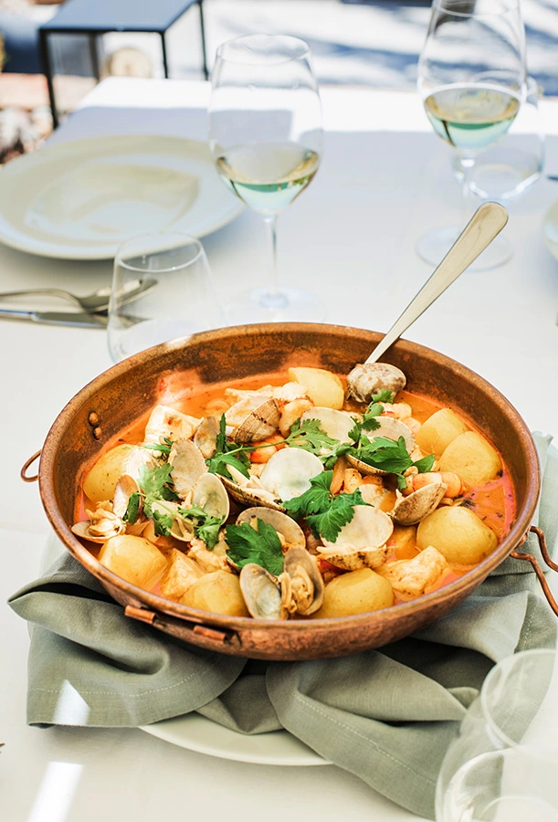Seafood stew with potatoes in a copper pot on a table with wine glasses.