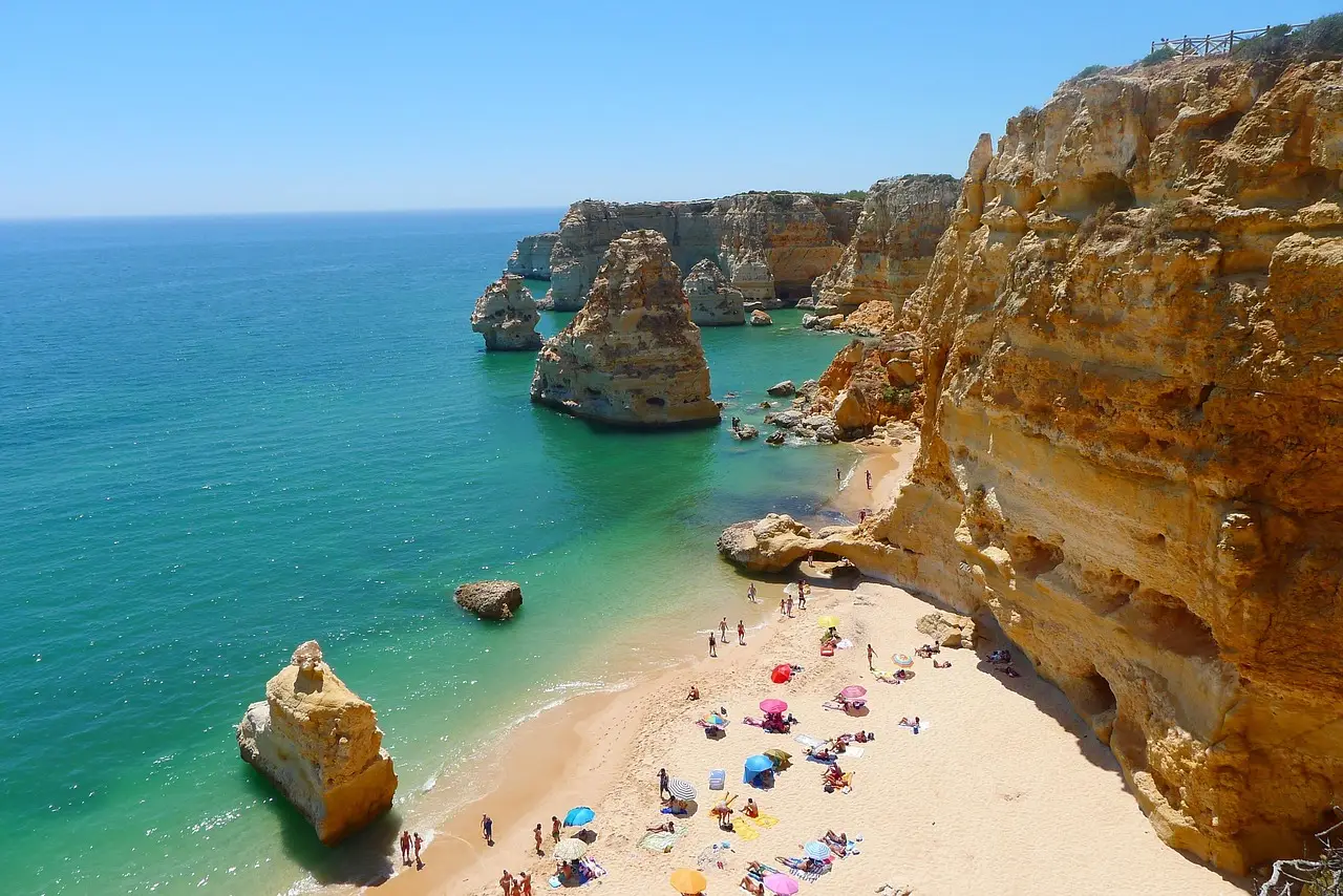 Sunny beach with cliffs, turquoise sea, and colorful umbrellas.