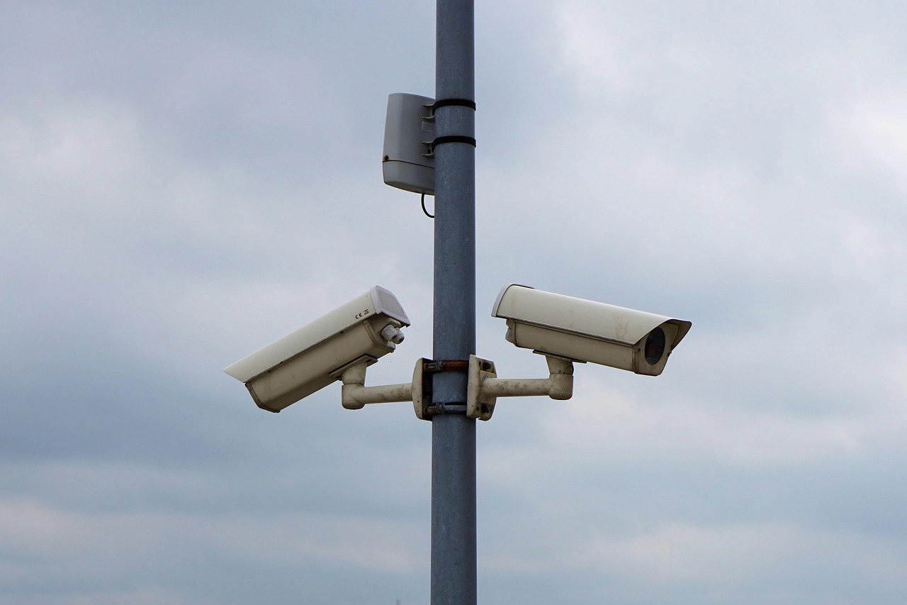 Two surveillance cameras mounted on a pole against a cloudy sky.