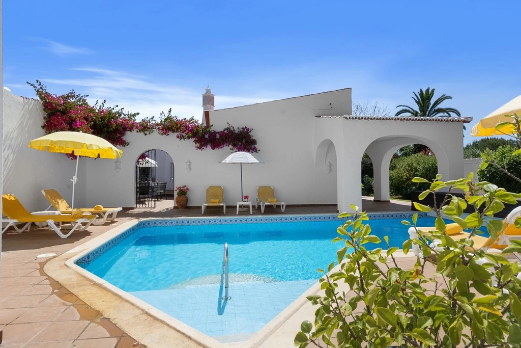 Sunny poolside with yellow loungers and umbrellas beside white villa walls.