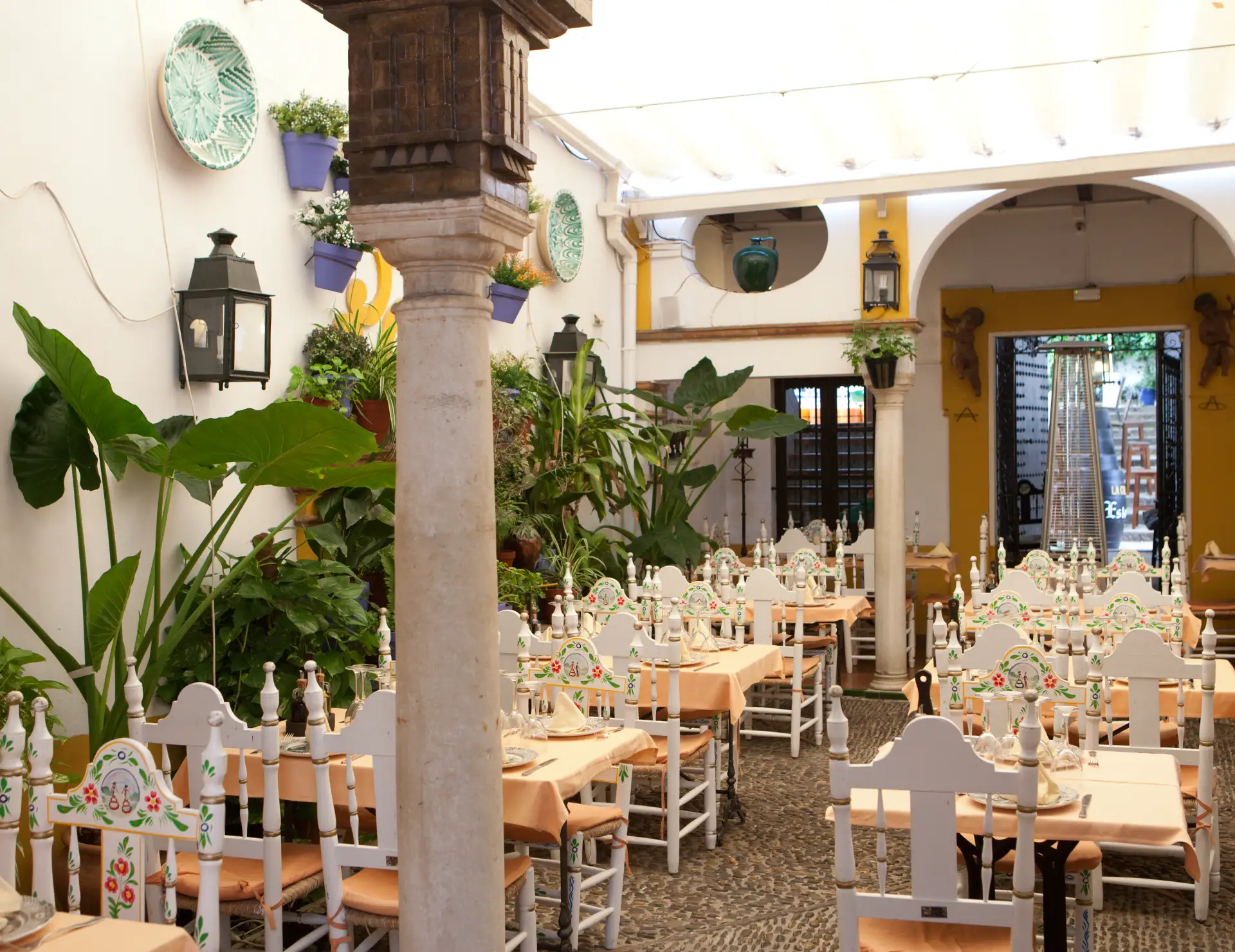 Patio con mesas y sillas decoradas, plantas y paredes blancas adornadas.
