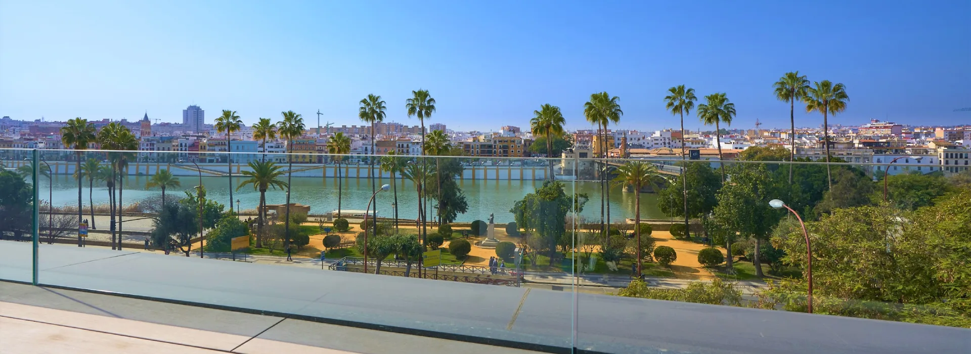 Vista de un r&iacute;o con palmeras y una ciudad al fondo bajo un cielo azul.