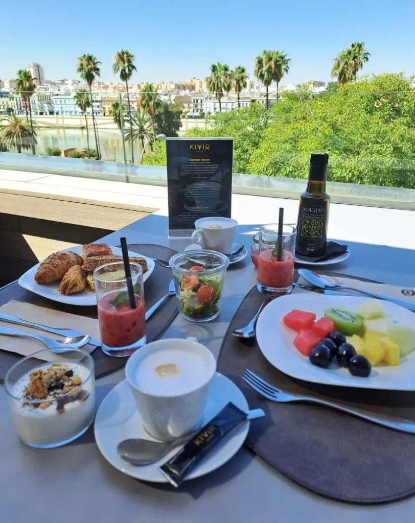 Desayuno al aire libre con fruta, pasteles y caf&eacute;, vista a palmeras y ciudad.