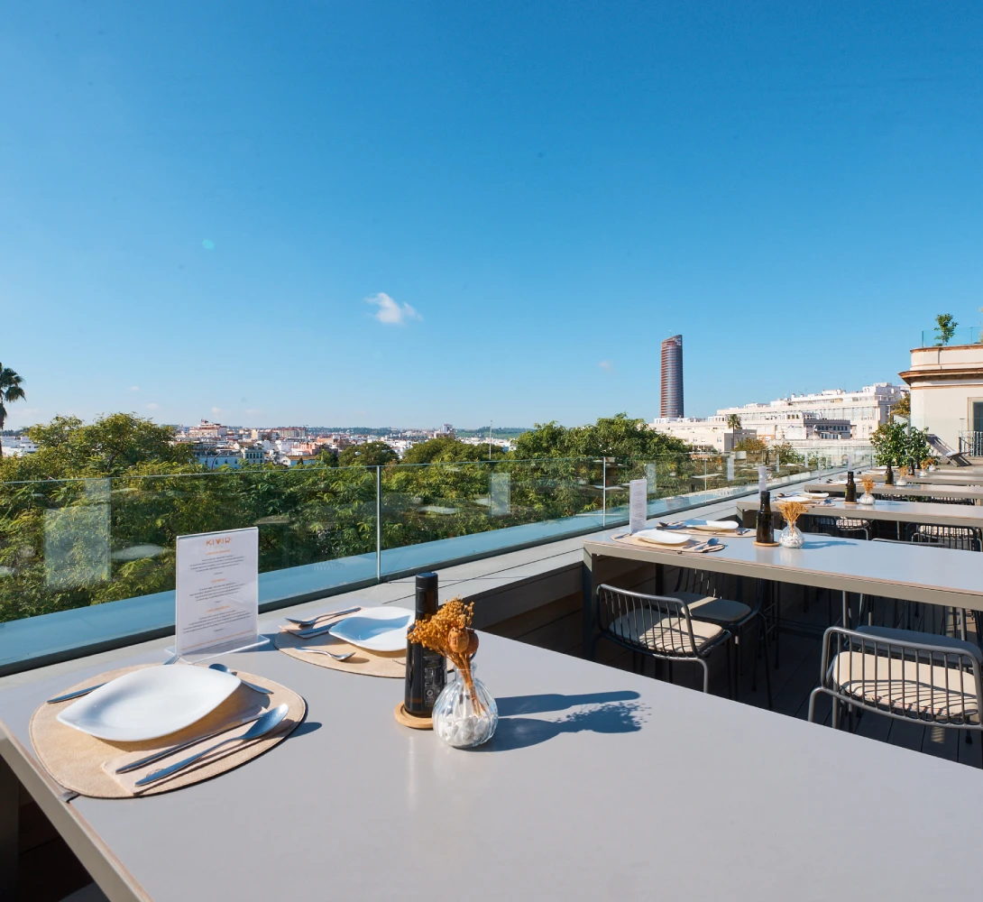 Terraza con mesas, sillas y vista de una ciudad bajo un cielo azul claro.