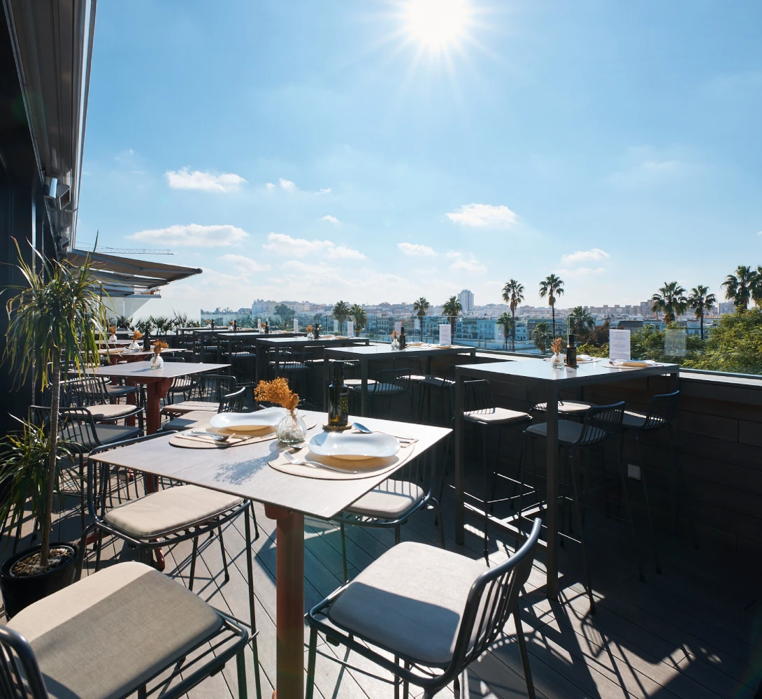 Terraza soleada con mesas y sillas, vista a la ciudad y palmeras.