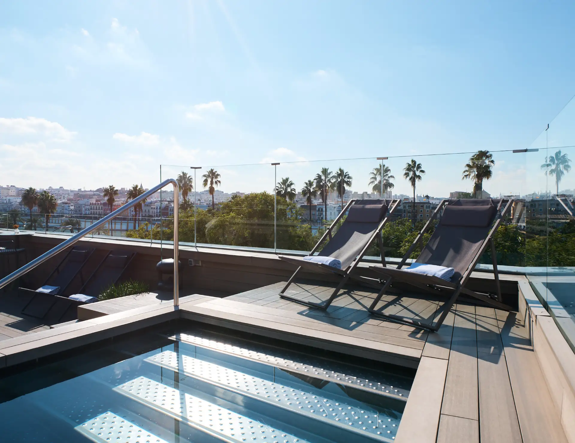 Terraza con tumbonas y piscina, vista de la ciudad y palmeras al fondo.