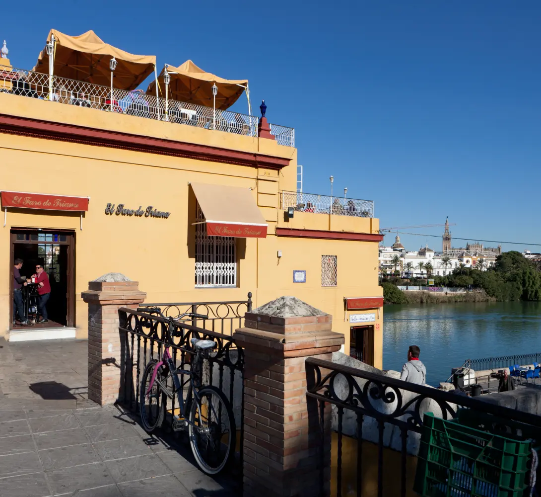Edificio amarillo junto al r&iacute;o con personas, bicicletas y terrazas al aire libre.