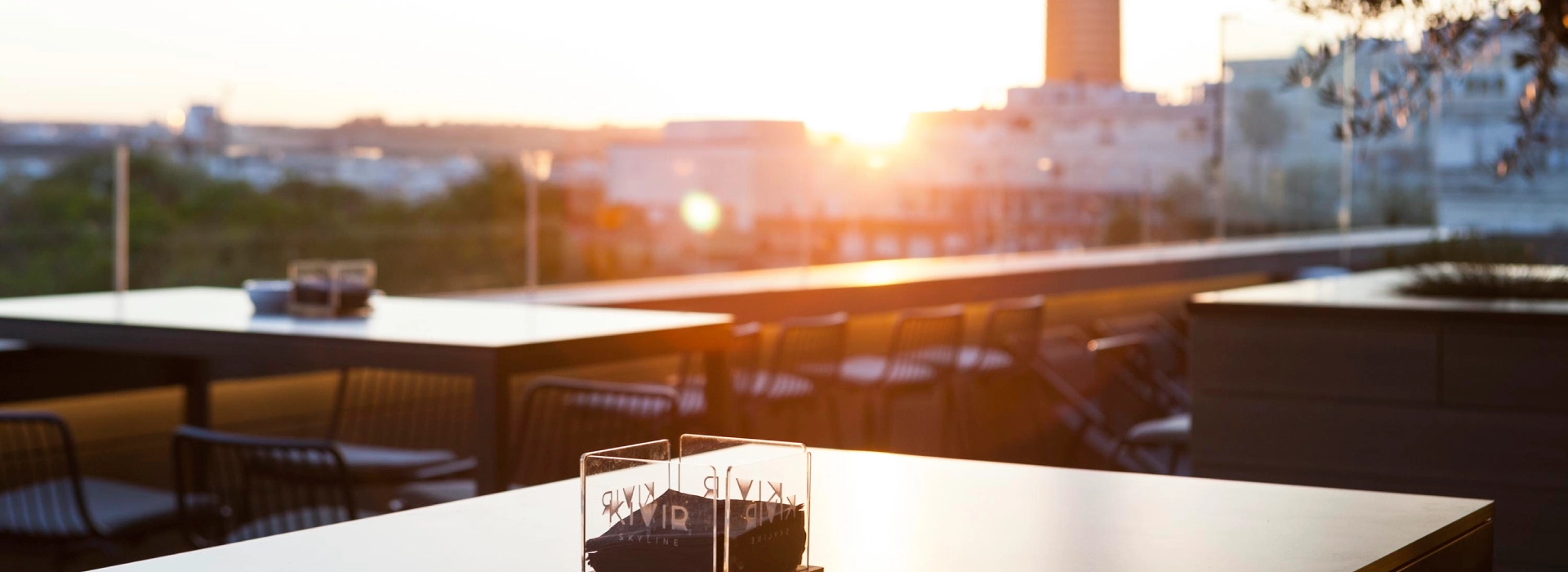 Terraza al atardecer con mesas y sillas vac&iacute;as, vista urbana de fondo.
