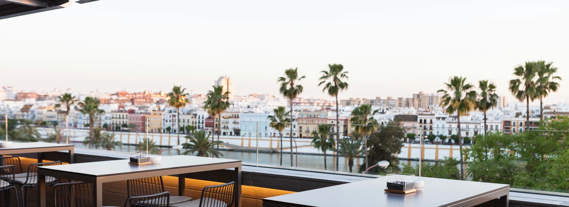 Terraza con mesas, sillas y vista panor&aacute;mica de una ciudad con palmeras.