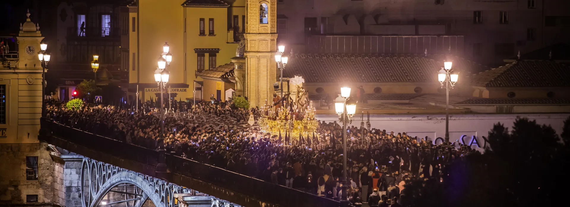 Procesi&oacute;n nocturna con multitud y luces en un puente.