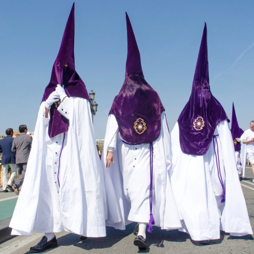 Tres personas con capirotes morados y t&uacute;nicas blancas en una procesi&oacute;n.