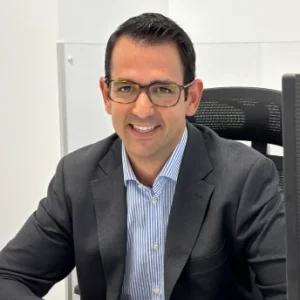 Hombre con gafas y saco sonr&iacute;e frente a una computadora en oficina.