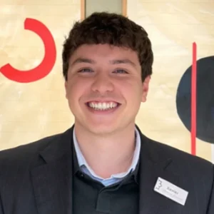 Joven sonriendo con saco y etiqueta de nombre sobre fondo art&iacute;stico geom&eacute;trico.