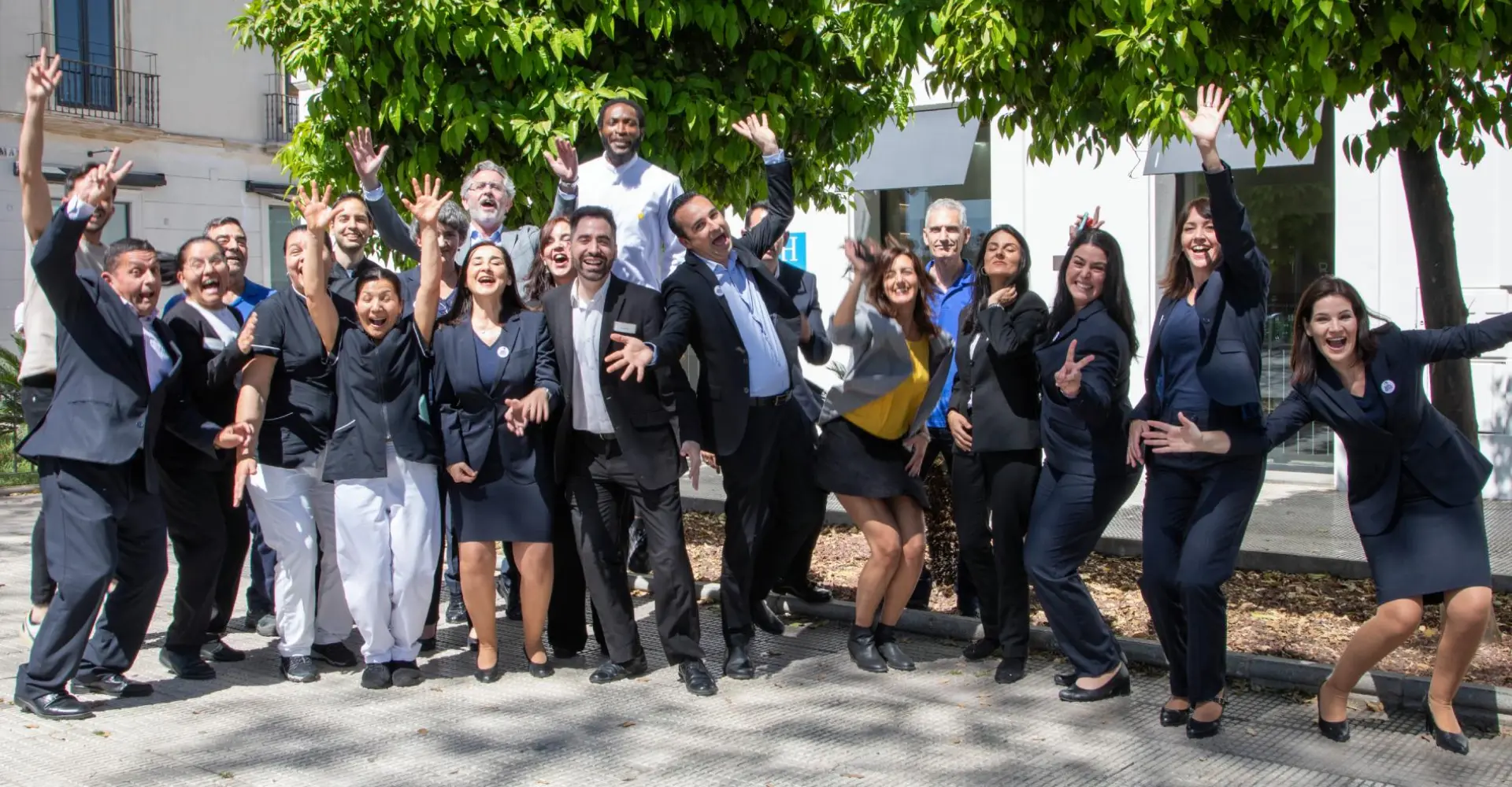 Equipo en traje, celebrando con manos alzadas frente a un edificio y &aacute;rboles