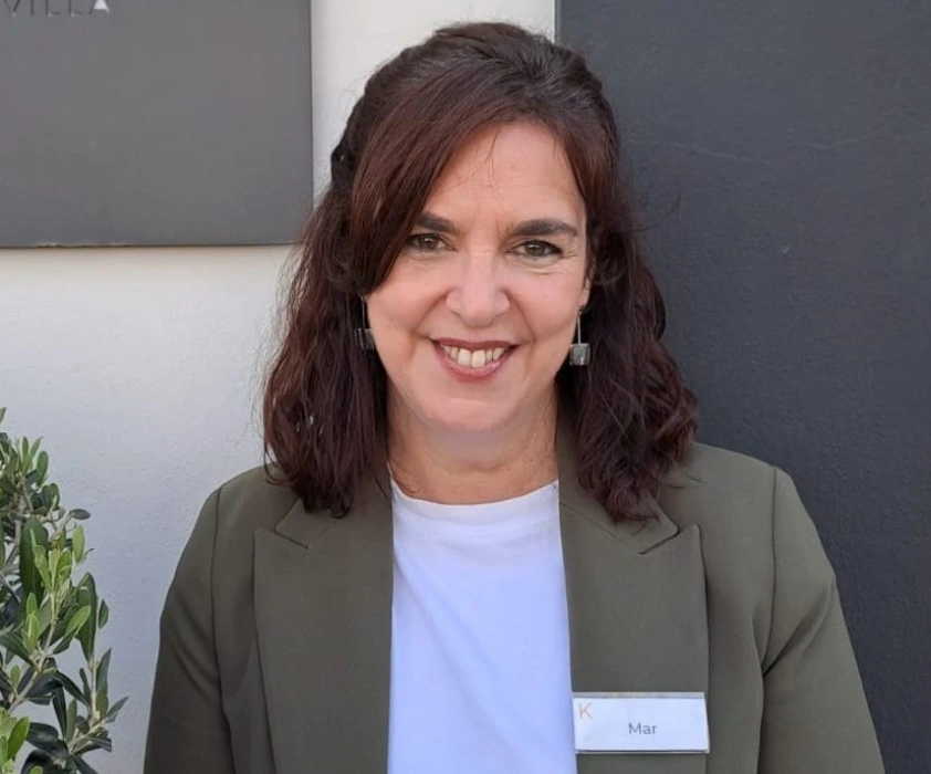 Mujer sonriendo vestida con saco verde y blusa blanca, lleva gafete de nombre.
