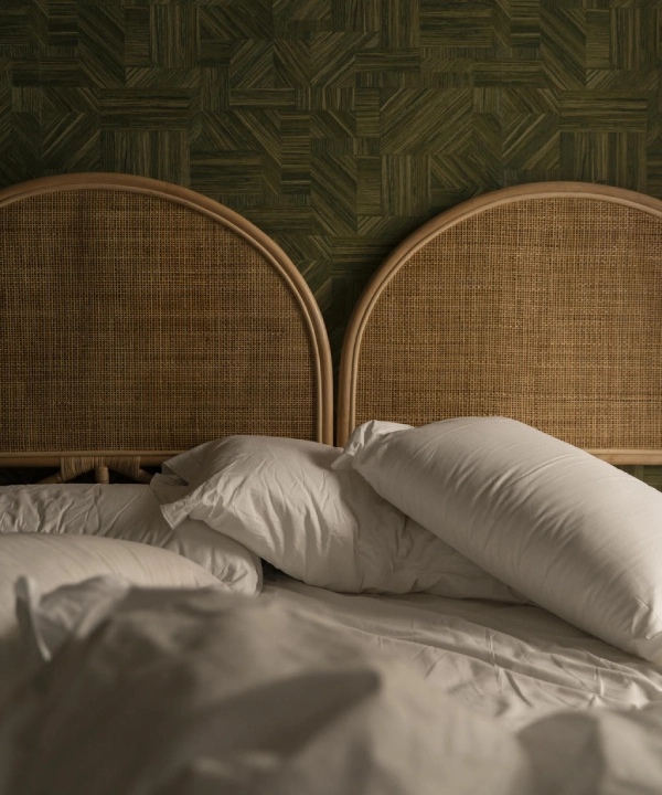 Cama desordenada con almohadas blancas y cabeceras de mimbre en fondo verde.