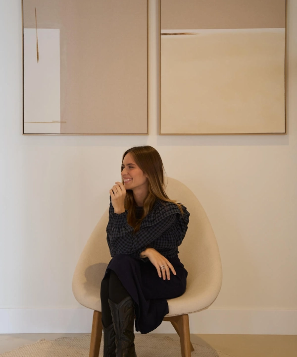 Mujer sonriendo sentada frente a dos cuadros abstractos en la pared.