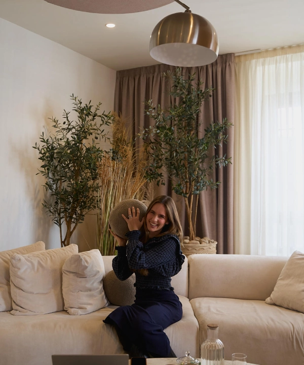 Mujer sonriendo en un sof&aacute; con cojines y plantas decorativas alrededor.