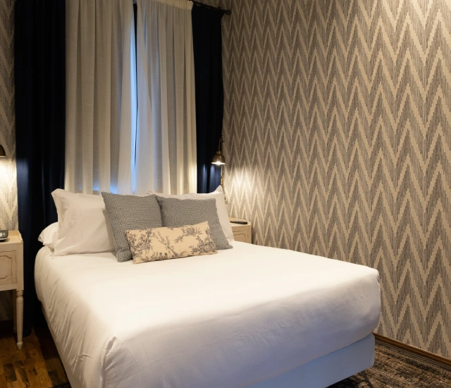 Cama con cojines en habitaci&oacute;n con papel tapiz de zigzag y cortinas oscuras.