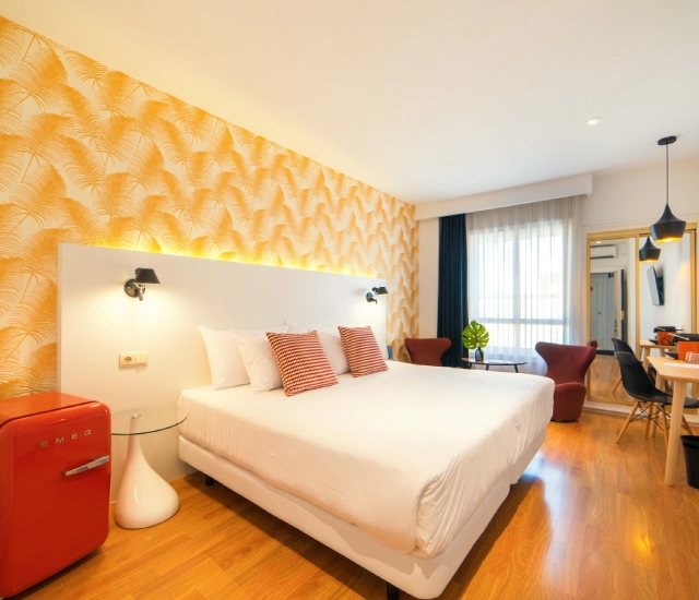 Habitaci&oacute;n moderna con cama, sillones, papel tapiz amarillo y mini nevera roja.