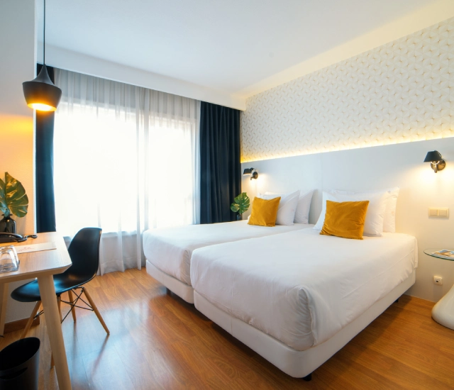 Habitaci&oacute;n moderna con dos camas, cojines amarillos y escritorio peque&ntilde;o.