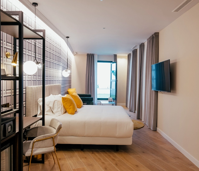 Moderna habitaci&oacute;n de hotel con cama, televisor y ventana al fondo.
