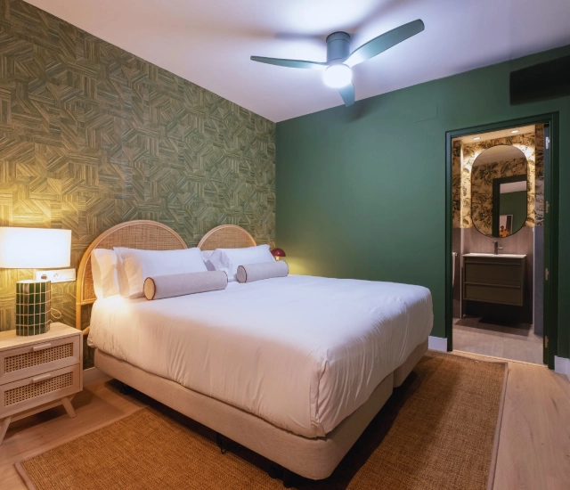 Habitaci&oacute;n moderna con cama doble, pared verde y ba&ntilde;o al fondo.