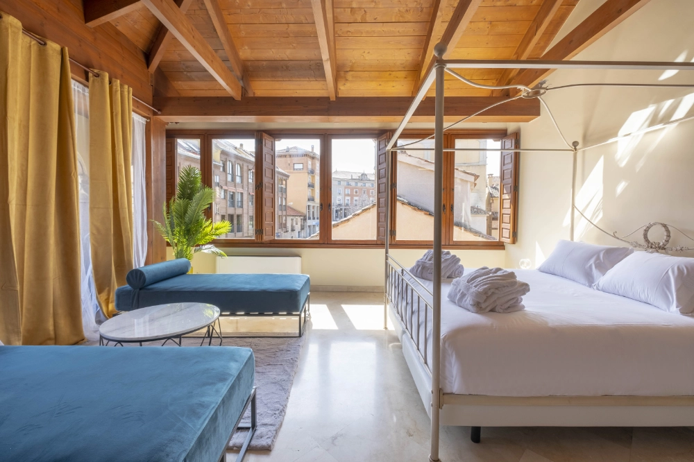 Habitación luminosa con cama, sofá azul, techos de madera y ventanales.