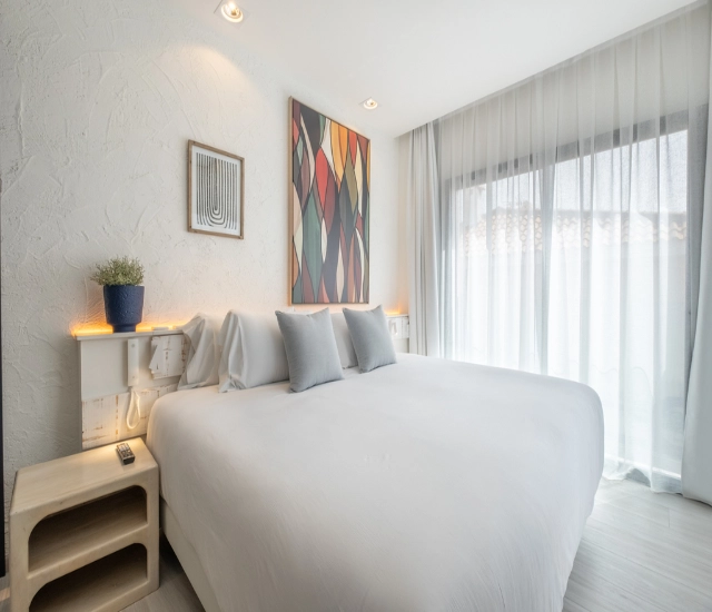 Habitaci&oacute;n luminosa con cama blanca, arte colorido y cortinas trasl&uacute;cidas.