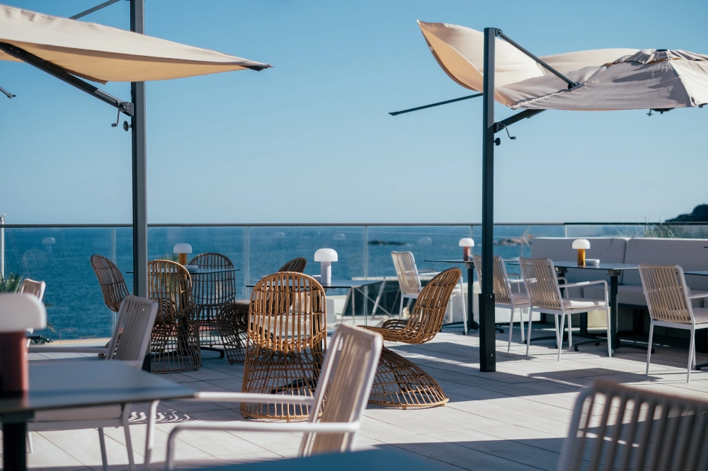 Terraza junto al mar con mesas, sillas de mimbre y sombrillas.