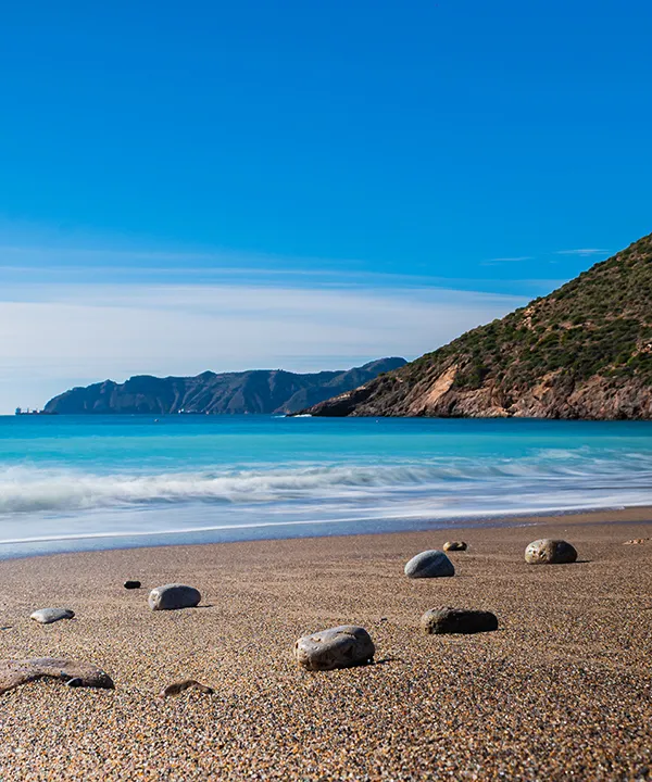 Playa con olas suaves, rocas y monta&ntilde;as al fondo bajo un cielo azul.